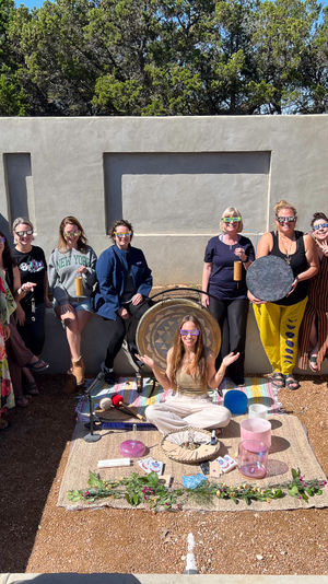 Sunlit outdoor stucco courtyard with about a dozen people wearing colorful eclipse glasses, gathered around a seated facilitator and a sound-healing setup with a gong, drum and crystal singing bowls on a woven mat, trees visible above the wall.