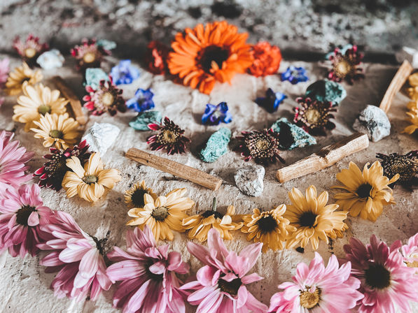 Boho flatlay of pink and yellow daisy blossoms, an orange gerbera, turquoise and silver stones and wooden sticks arranged on a rustic stone surface.