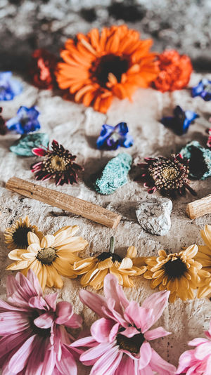 Boho flatlay of pink and yellow daisy blossoms, an orange gerbera, turquoise and silver stones and wooden sticks arranged on a rustic stone surface.