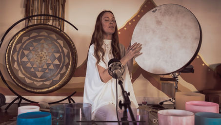 Indoor wellness studio sound healing: a woman with eyes closed plays a frame drum beside a gong and colorful crystal singing bowls, microphone in foreground.