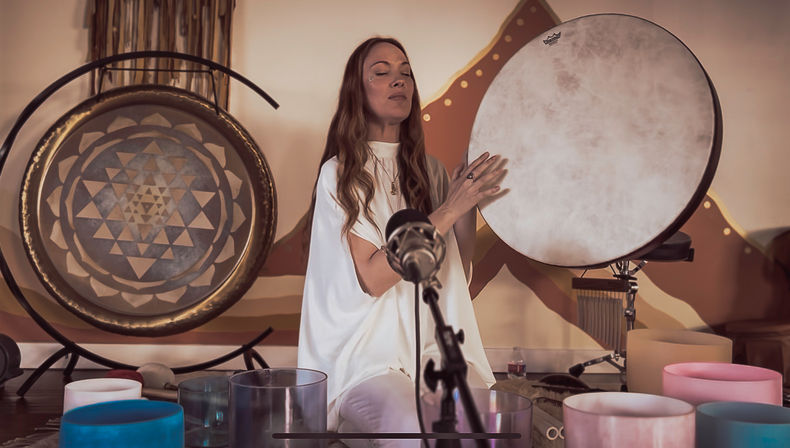Indoor wellness studio sound healing: a woman with eyes closed plays a frame drum beside a gong and colorful crystal singing bowls, microphone in foreground.