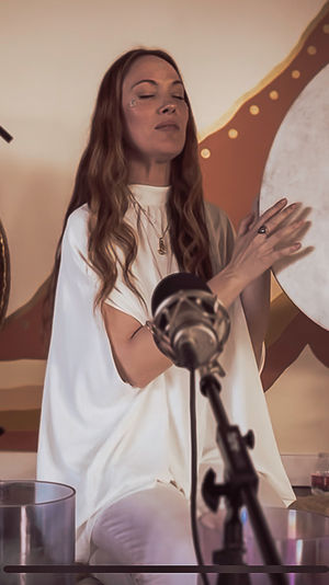 Indoor wellness studio sound healing: a woman with eyes closed plays a frame drum beside a gong and colorful crystal singing bowls, microphone in foreground.