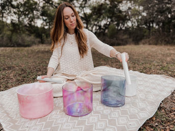 Person seated on a patterned blanket in a wooded outdoor setting, performing a sound bath and playing colorful crystal singing bowls with white mallets.