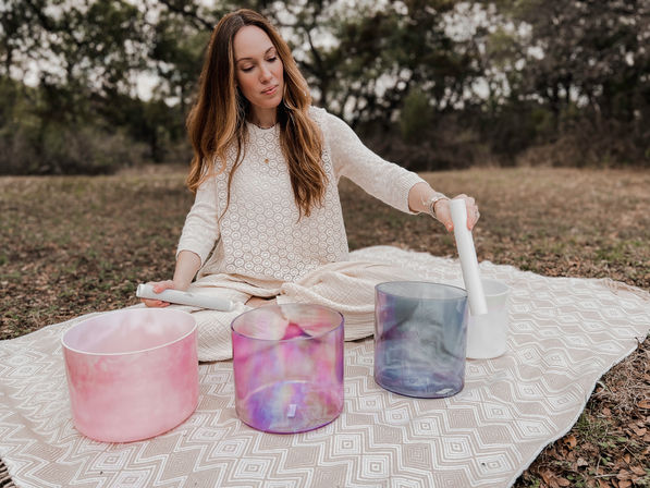 Person seated on a patterned blanket in a wooded outdoor setting, performing a sound bath and playing colorful crystal singing bowls with white mallets.
