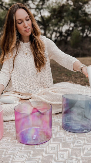Person seated on a patterned blanket in a wooded outdoor setting, performing a sound bath and playing colorful crystal singing bowls with white mallets.