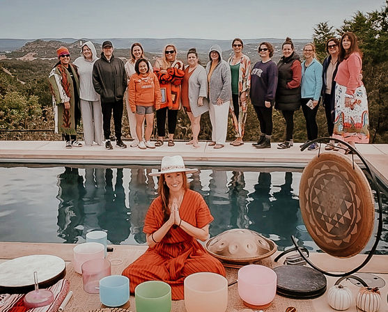 Hilltop wellness retreat by a pool: seated facilitator in a hat leads a sound-healing session with crystal singing bowls, handpan and gong while a group of guests stands along the pool edge against a scenic overlook.