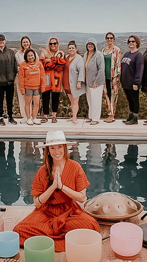 Hilltop wellness retreat by a pool: seated facilitator in a hat leads a sound-healing session with crystal singing bowls, handpan and gong while a group of guests stands along the pool edge against a scenic overlook.