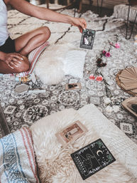 Hand holding a 'Transformation' tarot card over a boho patterned rug, surrounded by fluffy pillows, a tarot spread, crystals and pink flowers in a cozy living room setting.