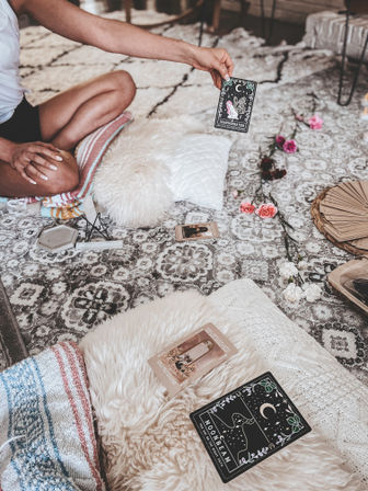 Hand holding a 'Transformation' tarot card over a boho patterned rug, surrounded by fluffy pillows, a tarot spread, crystals and pink flowers in a cozy living room setting.