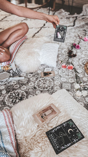 Hand holding a 'Transformation' tarot card over a boho patterned rug, surrounded by fluffy pillows, a tarot spread, crystals and pink flowers in a cozy living room setting.
