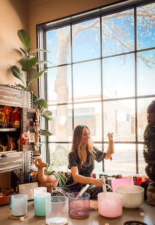 Sunlit urban storefront scene: a smiling woman seated by a large window in a wellness boutique playing a wooden chime and crystal singing bowls for a sound‑healing meditation session, surrounded by plants and carved wooden decor.