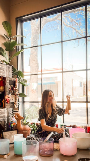 Sunlit urban storefront scene: a smiling woman seated by a large window in a wellness boutique playing a wooden chime and crystal singing bowls for a sound‑healing meditation session, surrounded by plants and carved wooden decor.
