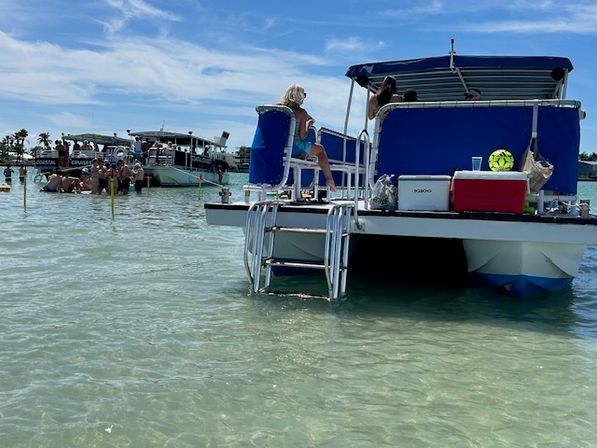Pontoon boat anchored in shallow clear turquoise water, passengers relaxing on blue seats with coolers and a swim ladder; sunny coastal scene with other boats and groups wading nearby.