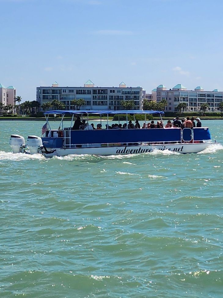 Blue-canopy pontoon tour boat full of passengers cruising turquoise coastal waters past beachfront condominiums under a sunny sky