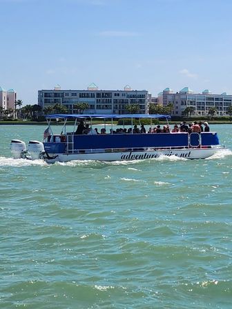 Blue-canopy pontoon tour boat full of passengers cruising turquoise coastal waters past beachfront condominiums under a sunny sky