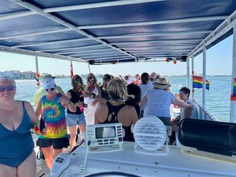Group of people enjoying a sunny party cruise on a covered pontoon boat in calm coastal waters, wearing tie-dye and leis with rainbow pride flags visible.
