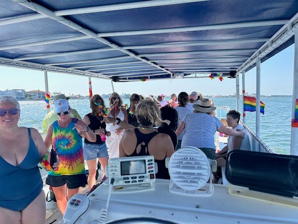 Group of people enjoying a sunny party cruise on a covered pontoon boat in calm coastal waters, wearing tie-dye and leis with rainbow pride flags visible.