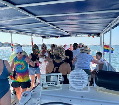 Group of people enjoying a sunny party cruise on a covered pontoon boat in calm coastal waters, wearing tie-dye and leis with rainbow pride flags visible.