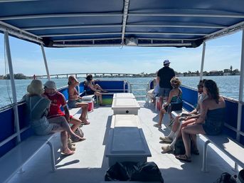 Passengers lounging on a covered pontoon boat cruising a calm blue bay, sunlit deck with bench seating and canopy, people in swimsuits and summer clothes, bridge and shoreline visible in the distance.