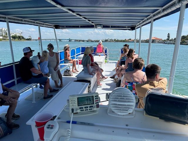 Relaxed passengers on a covered sightseeing boat tour cruising a sunny coastal waterway past a low bridge and palm-lined shoreline.