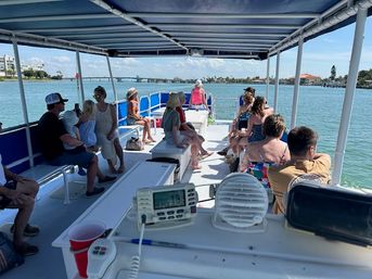 Relaxed passengers on a covered sightseeing boat tour cruising a sunny coastal waterway past a low bridge and palm-lined shoreline.
