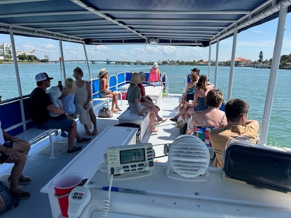Relaxed passengers on a covered sightseeing boat tour cruising a sunny coastal waterway past a low bridge and palm-lined shoreline.