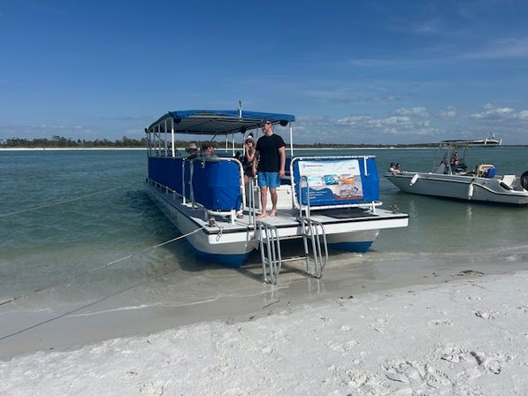Pontoon boat with blue canopy beached on white sandy shore, ladder down, people on board and a small powerboat nearby in turquoise coastal waters under a sunny blue sky.