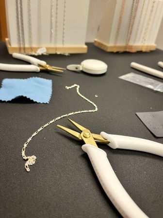 Close-up of a jewelry repair workspace with a delicate gold chain on a black mat, white-handled pliers and small precision tools, blue polishing cloth and blurred necklace displays in the background.