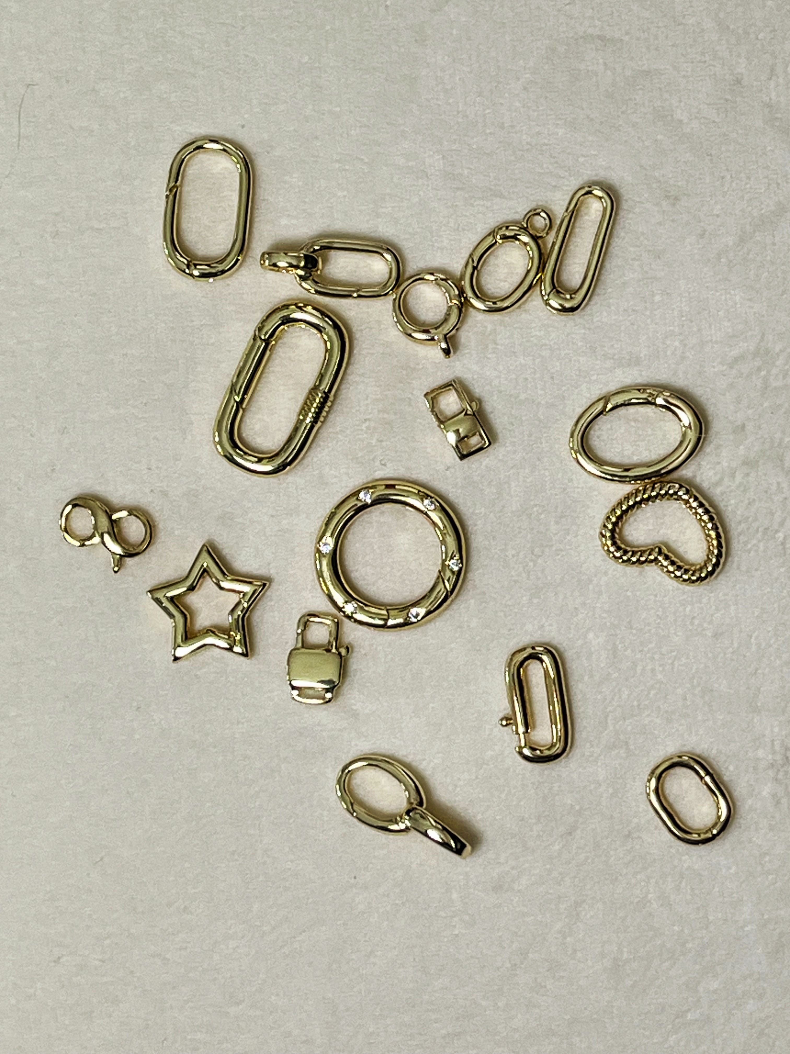 Flat-lay of scattered gold-tone jewelry findings and charms on beige fabric — oval links, round spring ring, lobster clasp, star and heart charms for DIY jewelry and accessories.