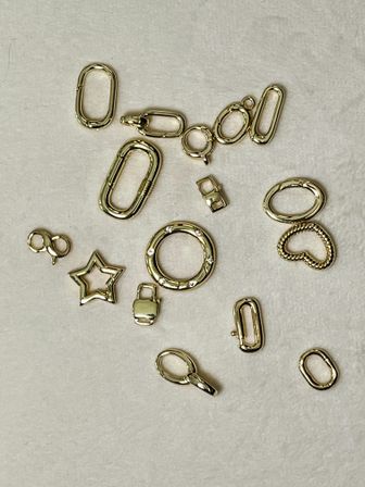 Flat-lay of scattered gold-tone jewelry findings and charms on beige fabric — oval links, round spring ring, lobster clasp, star and heart charms for DIY jewelry and accessories.