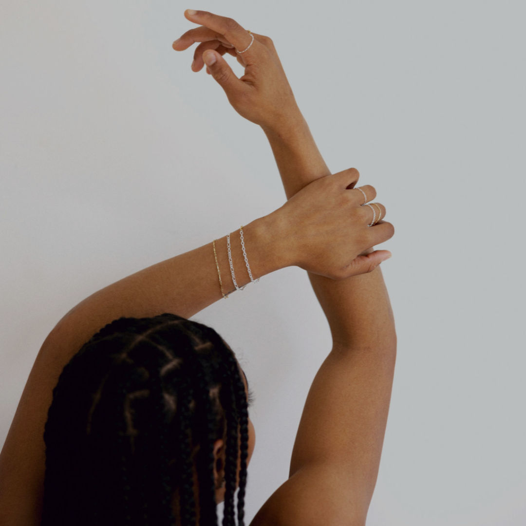Close-up of raised arms with braided hair showcasing delicate gold and silver chain bracelets and stacking rings against a minimal white studio background.