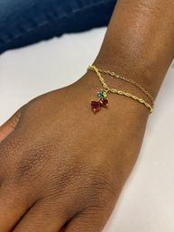 Close-up of a dark brown wrist wearing two delicate gold chain bracelets; one features a sparkling red heart-shaped gemstone with a small green leaf accent pendant on a white background.