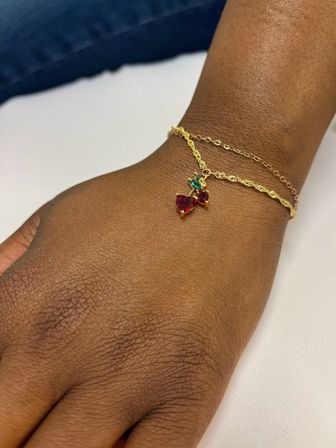 Close-up of a dark brown wrist wearing two delicate gold chain bracelets; one features a sparkling red heart-shaped gemstone with a small green leaf accent pendant on a white background.