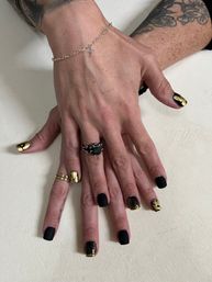 Close-up manicure: hands with black matte and gold-foil nail art, rings and silver cross bracelet, floral tattooed forearm on neutral background