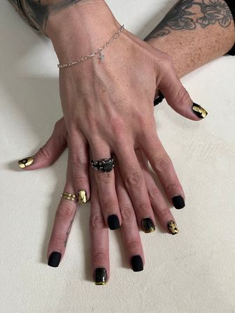 Close-up manicure: hands with black matte and gold-foil nail art, rings and silver cross bracelet, floral tattooed forearm on neutral background