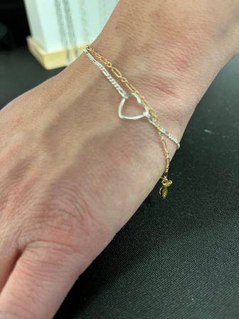 Close-up of a wrist wearing a chic minimalist layered bracelet stack: a silver open-heart charm paired with a thin gold chain and tiny gold charm on a black background