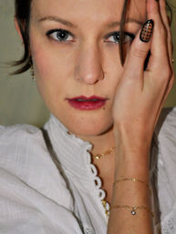 Indoor close-up portrait of a woman with a striking gaze, smoky eye makeup and dark red lipstick, hand partly covering one side of her face revealing a long polka-dot nail, wearing a white ruffled blouse and delicate gold bracelets and necklace.