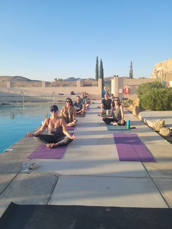 Outdoor group yoga class on mats along a pool deck at sunset, people meditating beside a calm pool with desert hills and cypress trees under a clear blue sky.