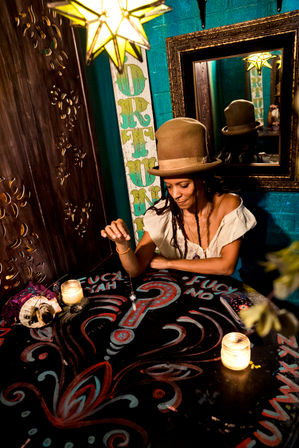 Fortune-teller in a brown hat doing a pendulum reading over a painted bohemian table with lit candles, star lantern, ornate mirror and teal-accented walls.