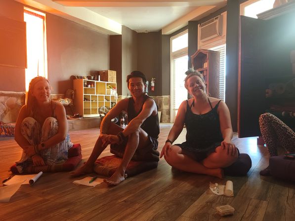 Three smiling adults sitting cross-legged on floor cushions in a sunlit indoor yoga/meditation studio with wooden floors and cozy shelving