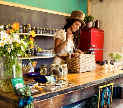 Woman in a hat arranging a woven basket on a rustic wooden counter inside a cozy herbal apothecary shop with mason jars of dried herbs, fresh daisies, candles and a red retro fridge.