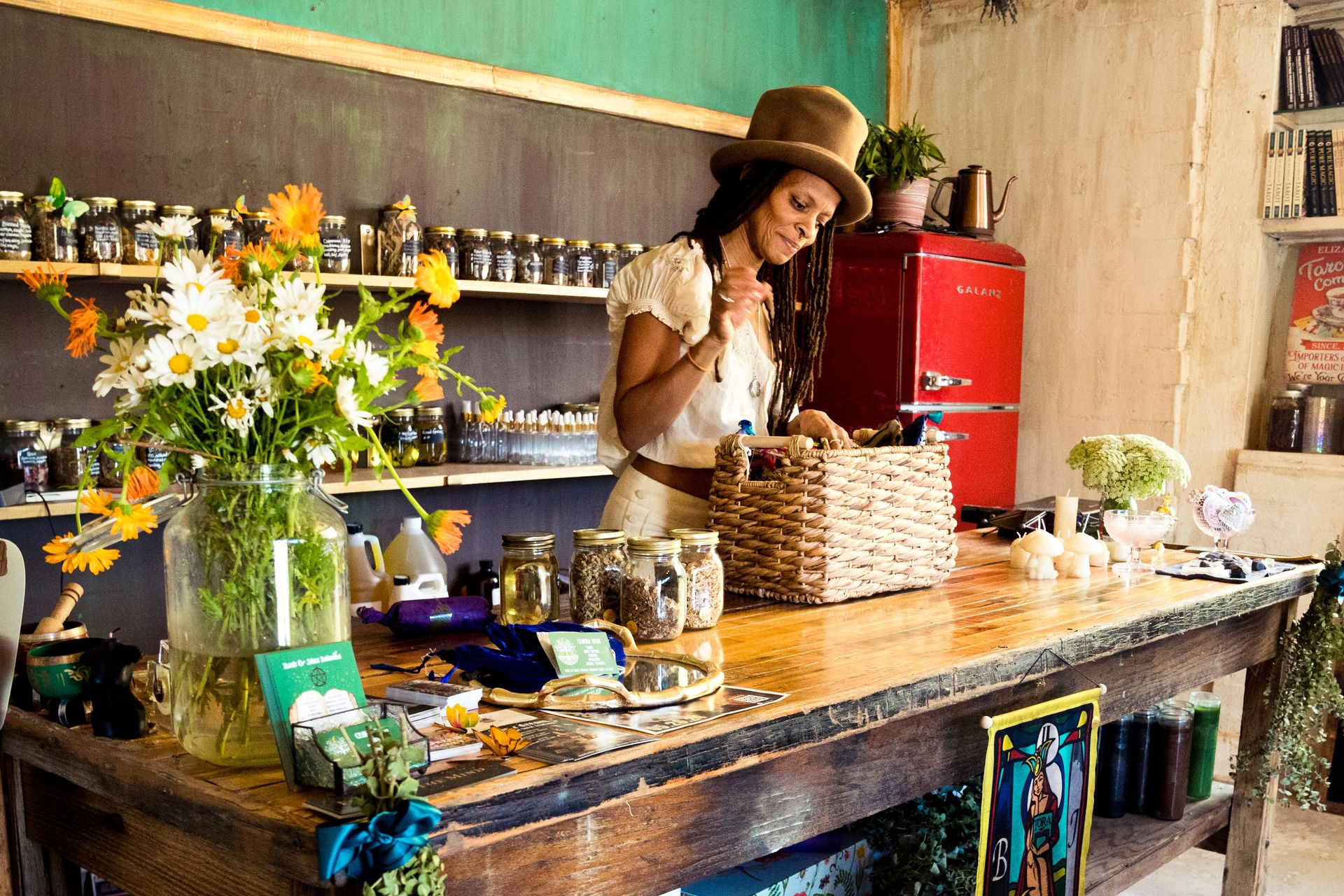 Woman in a hat arranging a woven basket on a rustic wooden counter inside a cozy herbal apothecary shop with mason jars of dried herbs, fresh daisies, candles and a red retro fridge.