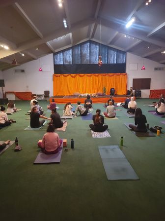 Group yoga class in a spacious community hall — students seated on mats on a green carpet facing an orange-draped stage with cushions and soft overhead lighting.