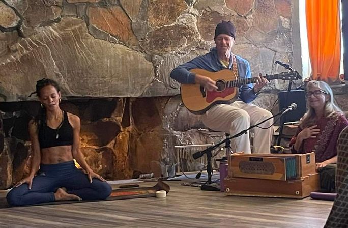 Woman in seated yoga pose on a mat beside a guitarist and harmonium player in a cozy studio with a large stone fireplace — indoor yoga and live acoustic music session for wellness and meditation.