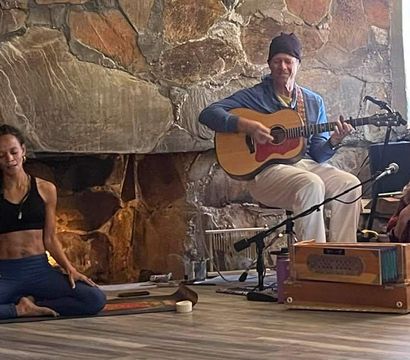 Woman in seated yoga pose on a mat beside a guitarist and harmonium player in a cozy studio with a large stone fireplace — indoor yoga and live acoustic music session for wellness and meditation.