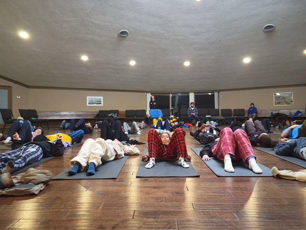 Group relaxation session on yoga mats in a hardwood floor community room, participants lying on their backs in cozy pajamas and casual clothes under warm ceiling lights.