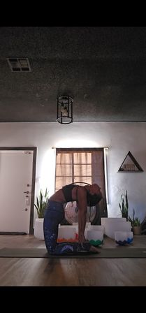 Person in camel yoga pose on a mat in a sunlit wellness studio with crystal singing bowls, a gong, potted plants and galaxy-print leggings.