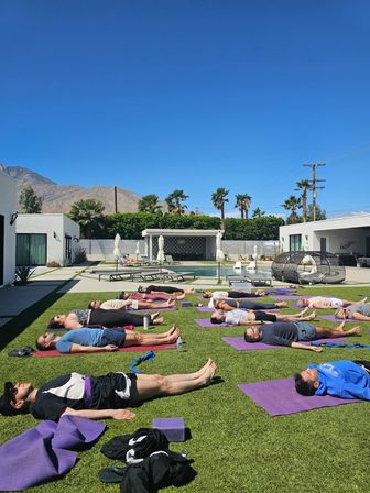 Poolside outdoor yoga class at a sunny desert retreat — people in savasana on purple mats on green lawn, modern white villas, palm trees and mountains under a clear blue sky