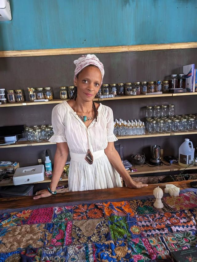 Woman in a white dress and headscarf standing behind a wooden counter in a boho artisan apothecary-style shop, shelves lined with glass jars and bottles, colorful embroidered patchwork cloth and candles on the counter.