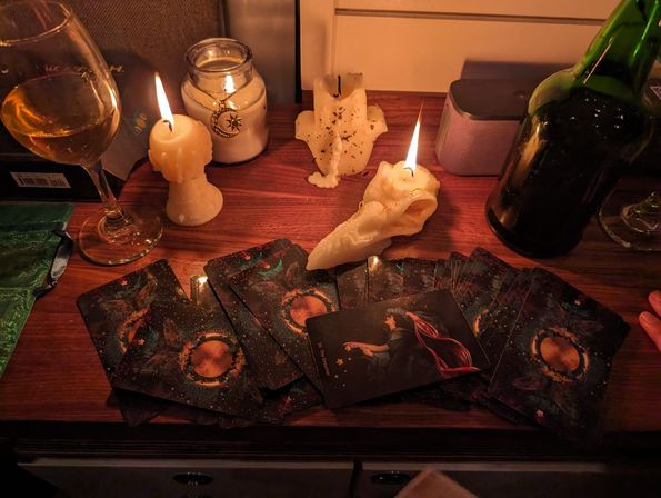 Candlelit tarot reading on a wooden tabletop: cosmic-themed tarot cards fanned out amid melted wax candles, a jar with moon charm, plus a wine glass and green bottle.